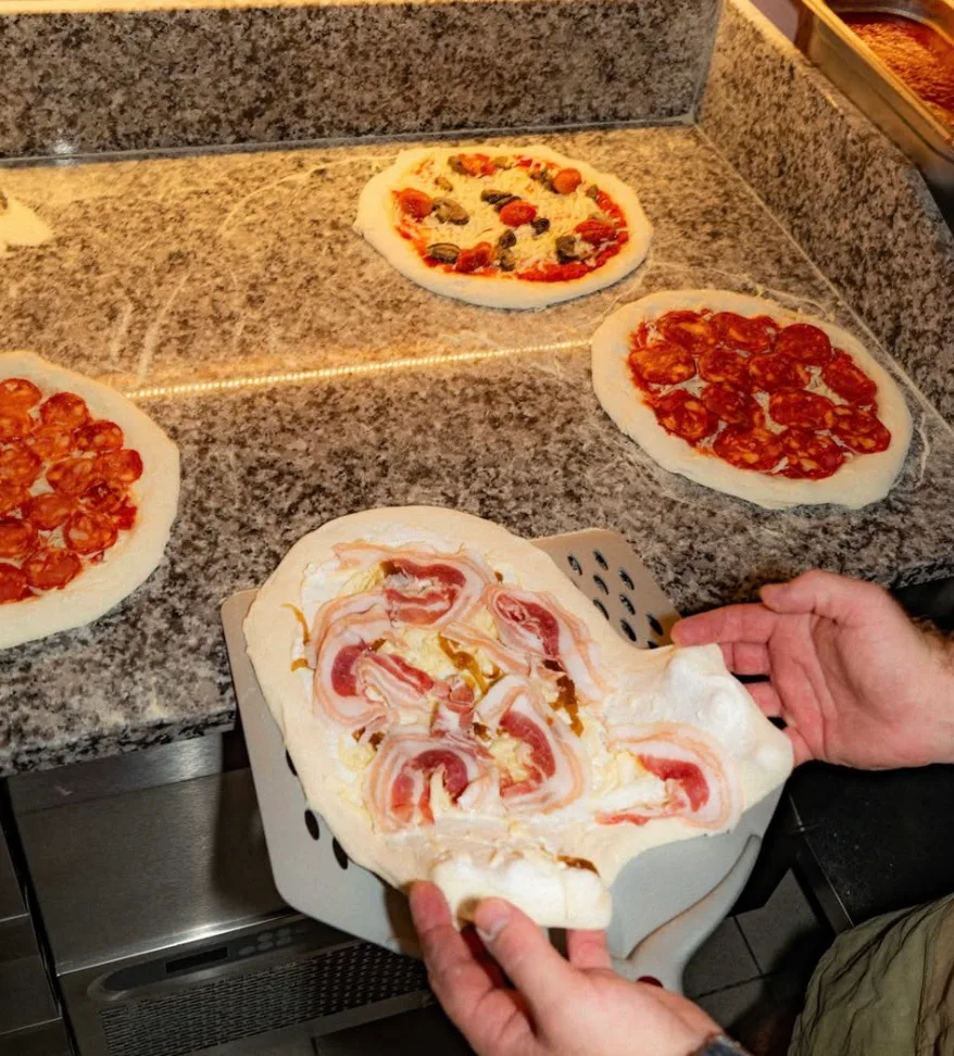 Hands preparing unbaked pizzas with various toppings at Ipanema bar in Kaunas.