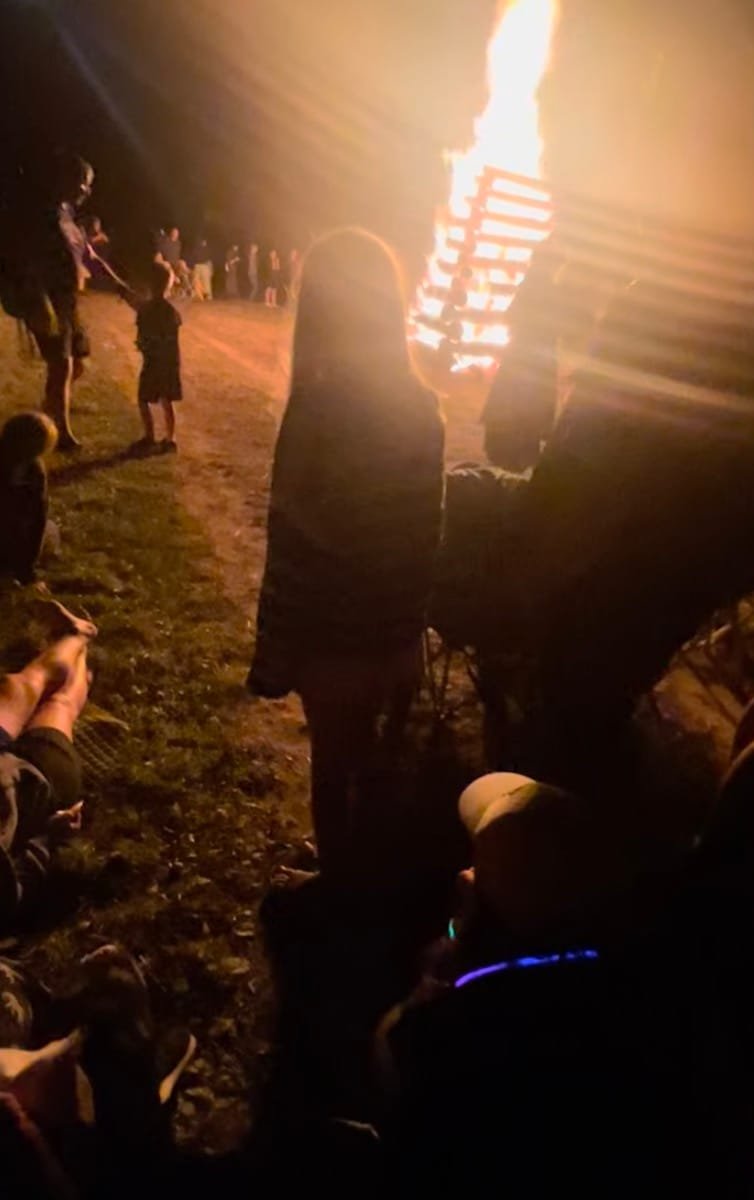 People gathered around a tall Joninės Midsummer bonfire at night.