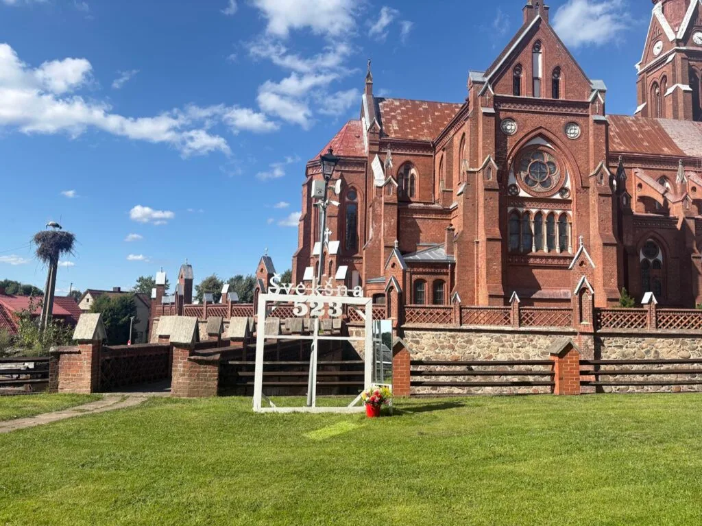 Red brick Neo-Gothic church in Švėkšna, Lithuania, with decorative town sign on green lawn