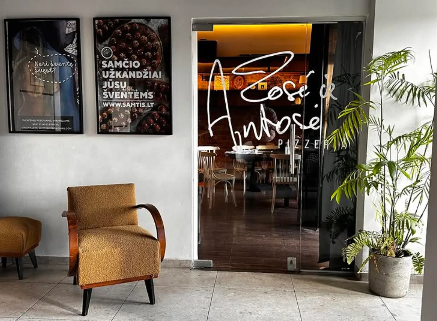 Entrance area with armchair and glass door leading into Žosė ir Antosė pizzeria.