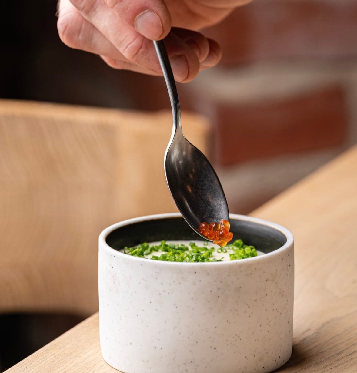 A hand adding red caviar with a spoon onto a gourmet dish served in a ceramic bowl.
