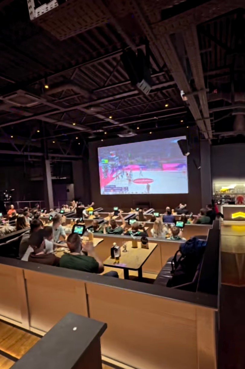 Crowd watching a sports game on a large screen at Action! by Apollo sports bar.