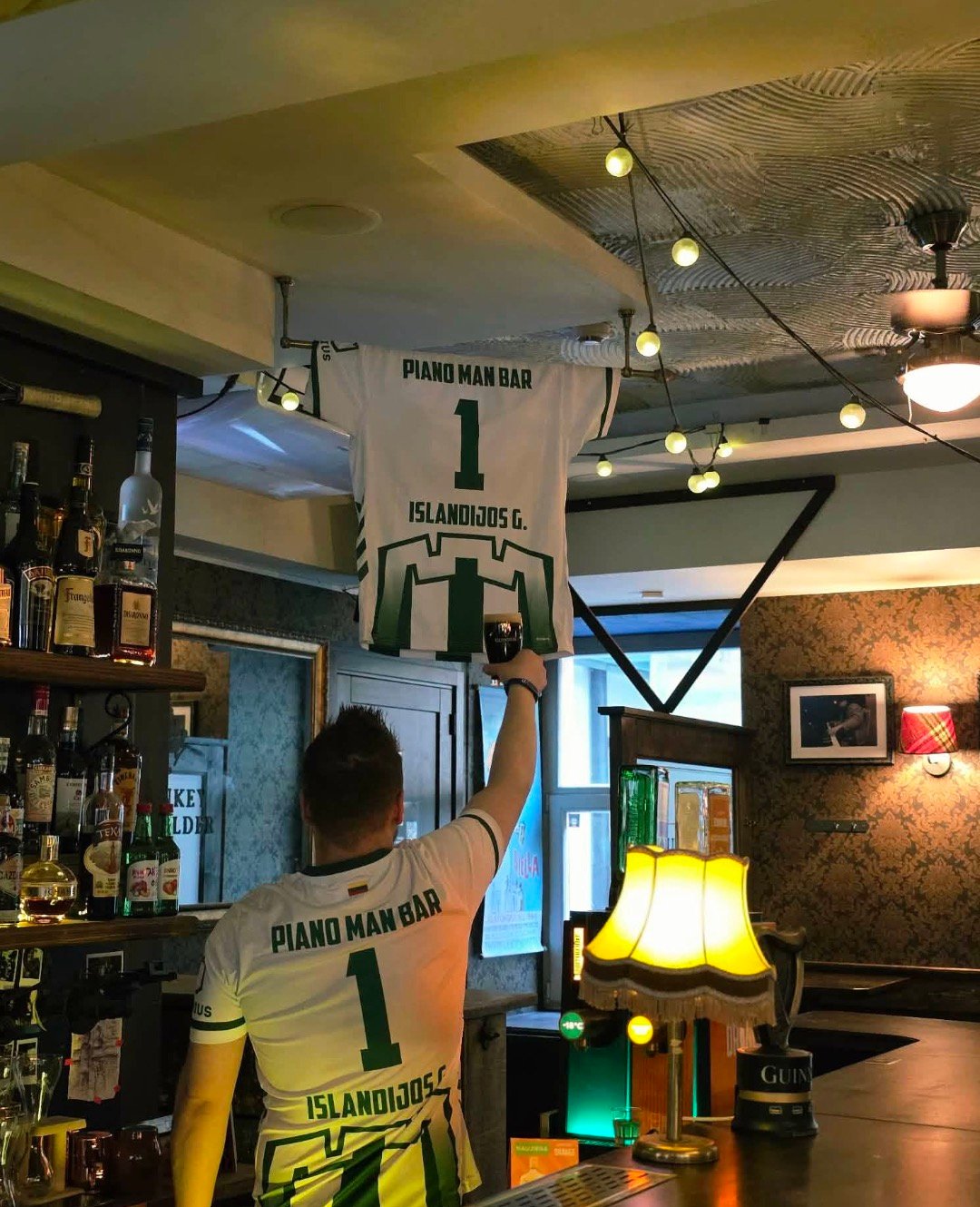 Man holding up a beer glass under a Piano Man Bar jersey inside the pub.