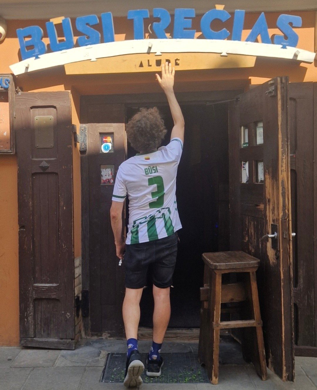 Man in sports jersey waving at the entrance of Būsi Trečias pub in Vilnius.
