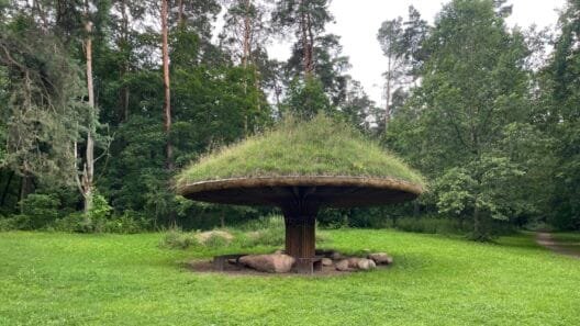 A wooden mushroom-shaped pavilion covered in grass sitting on a sunny forest lawn