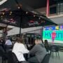 People sitting at outdoor tables watching sports on a large screen at sports bars in Vilnius